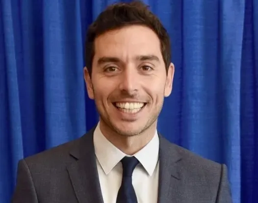 Headshot of Dr. Marc Oliva smiling facing forward. He is wearing a dark-gray suit and has short dark-brown hair. He is in front of a blue curtain background.