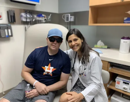 Dr. Ferrarotto with a patient in clinic. Dr. Ferrarotto is wearing a white coat and stethoscope, and is smiling facing forward. She is next to her patient, who is wearing black sunglasses and a dark blue shirt.