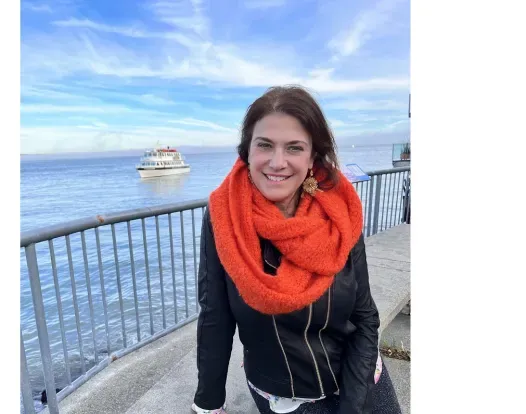 Lillian Kreppel wearing an orange scarf and a black long-sleeved outfit. She is smiling facing forward, sitting on a ledge nearby a bay. The sea and sky merge on the horizon and it is sunny and blue.
