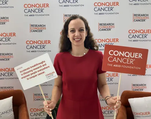 Headshot of Dr. Lauren Byers at the ASCO Annual Meeting. She's holding a sign that says Conquer Cancer, the ASCO Foundation, against a backdrop with tiles that say Research Conquers Cancer.