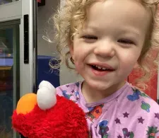 Leo with medium-length curly hair, smiling and laughing, while holding an Elmo plushie.