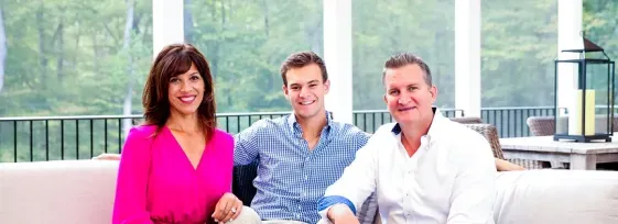 Left to right: Nancy Berry, Jake Berry, Alex Berry. All three are seated facing forward and smiling, located in a screened-in sun-room with white columns and green trees visible in the background. Nancy is wearing a bright pink shirt with dark blue pants. Jake is wearing a light blue shirt with khaki plants. Alex is wearing a white shirt with black pants.