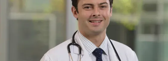Dr. Michael Kinnaman in a doctor's coat with a stethoscope, smiling facing forward in an outdoor setting