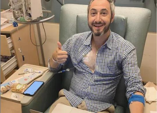 Dave Thau in a clinical setting, smiling and holding a thumbs-up. He has a monitor attached to his chest and is sitting, with a laptop.