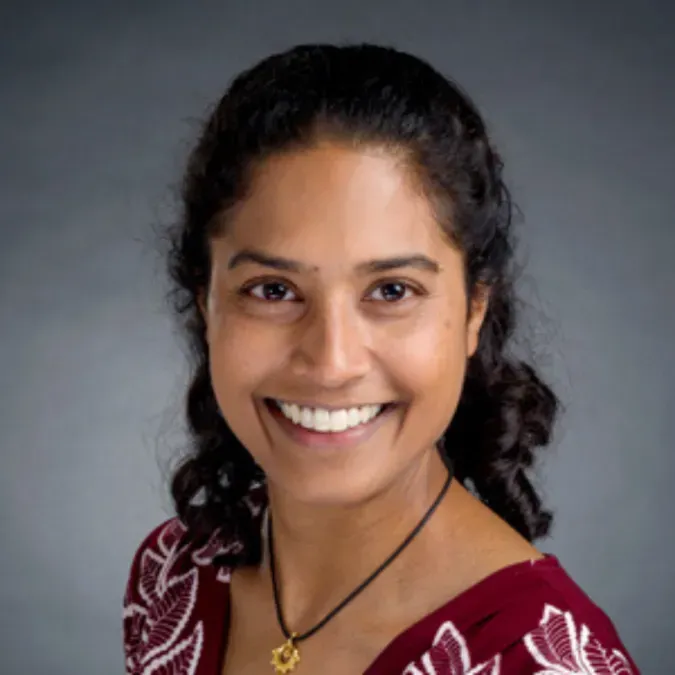 Dr. Sheena Mukkada facing forward and smiling. She has shoulder-length black hair and is wearing a dark-red shirt with a black necklace, against a plain gray background.